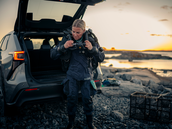 Person i dykkerudstyr foran en Subaru Solterra med åben bagklap ved havet.