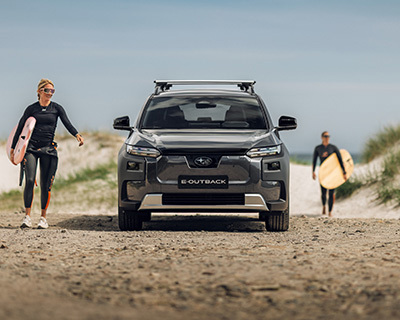 Subaru E-Outback på en strandvej med to personer, der bærer surfbrætter.