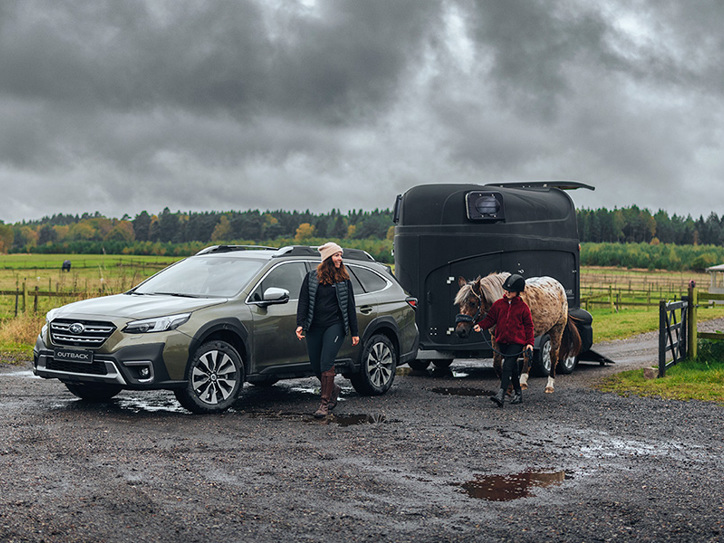 Subaru Outback med hestetrailer i mudder, mens en kvinde og en pige går ved siden af – pigen fører en hest.
